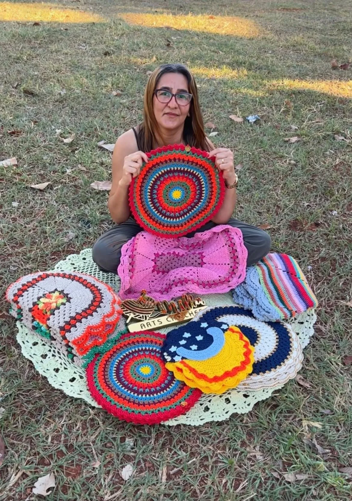 Milla sitting outdoors on the grass, holding a round crocheted mandala, surrounded by her pieces and a plaque reading Milla Arts Crochês