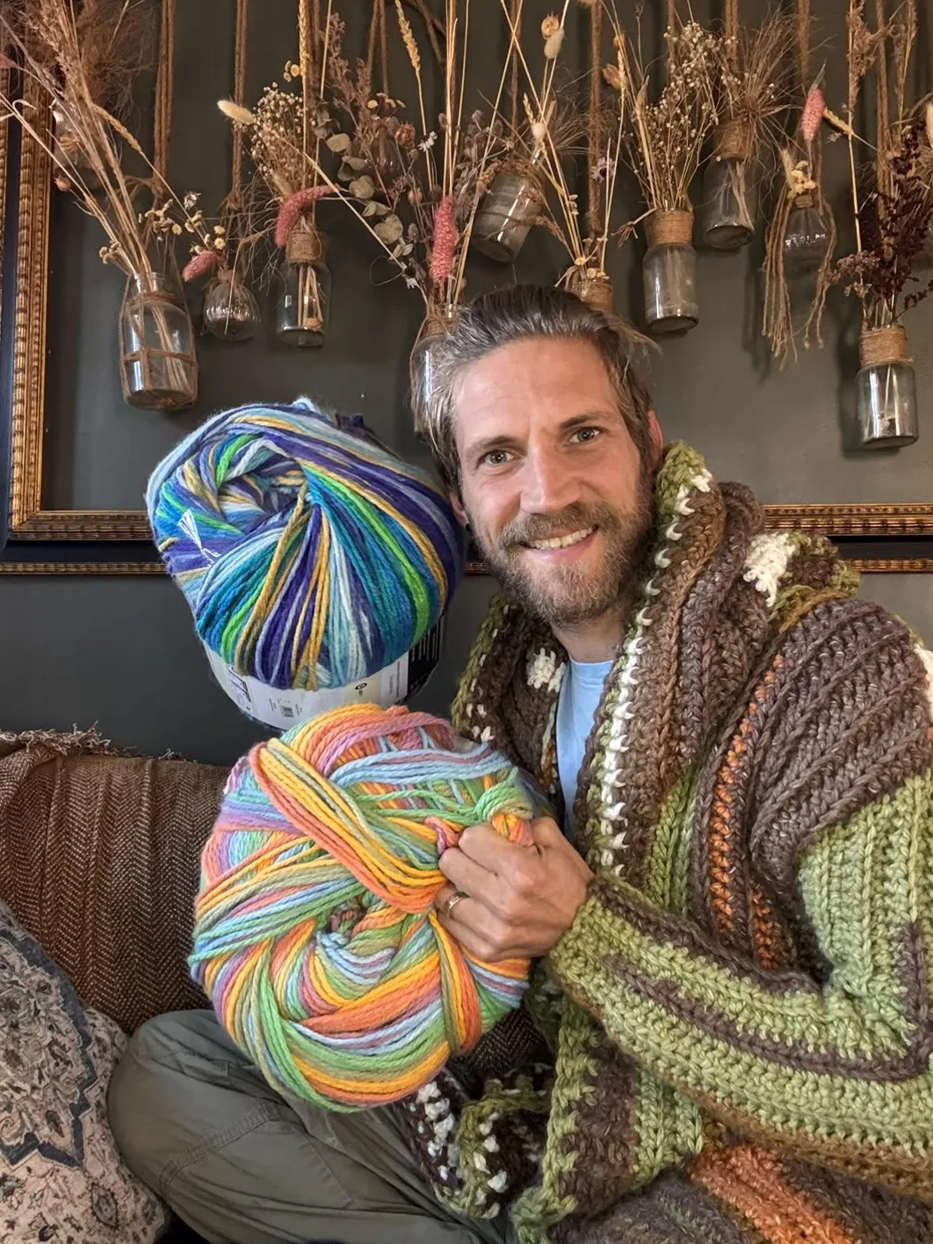 Vic smiling on a sofa in a hooded crocheted cardigan, holding two large balls of multicoloured yarn